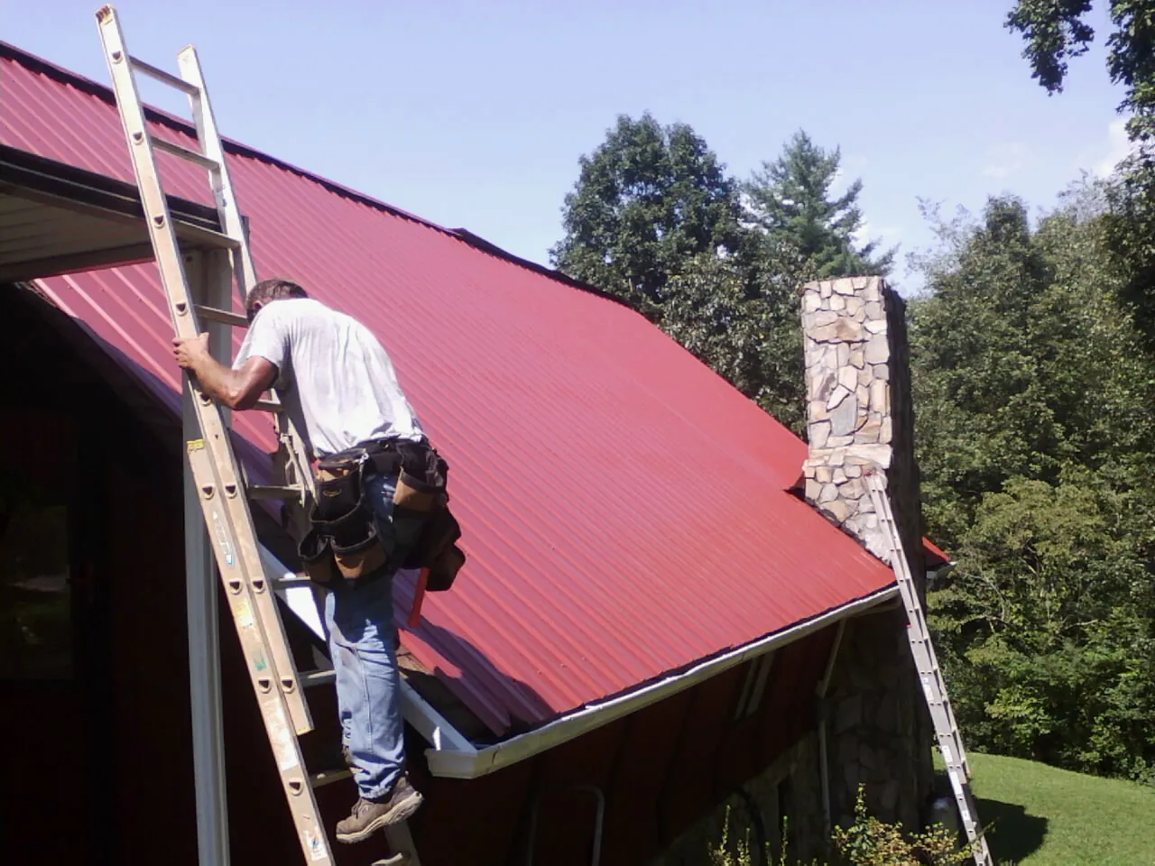 Roofer on a ladder beside a red metal roof and stone chimney — Soffit & Fascia Repair by Roofing Brothers in Fulks Run